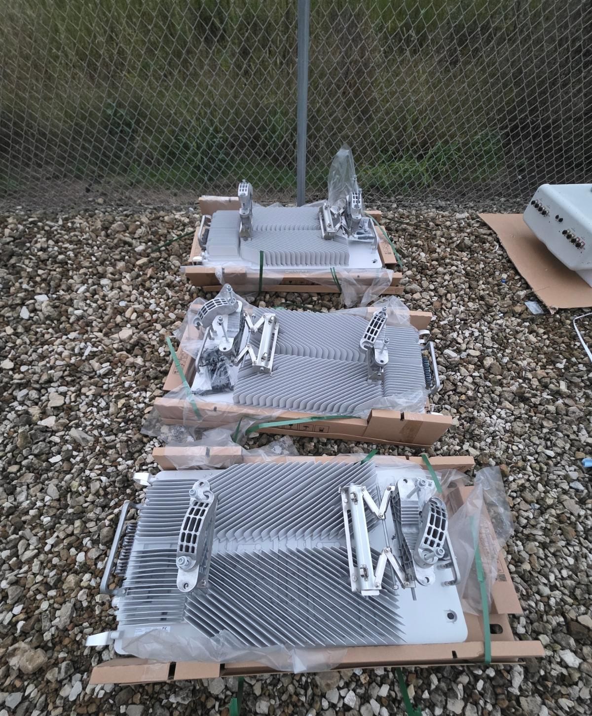 Three radios with mounting brackets attached, on their shipping crates on the gravel compound inside the fence.