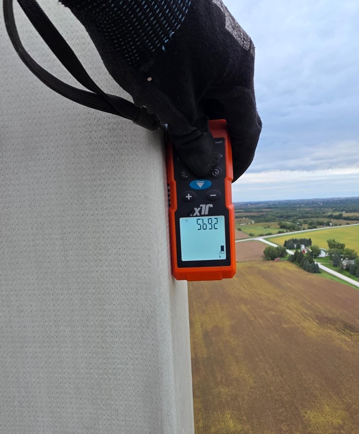 A gloved hand holds an orange laser measure against an antenna at the top of the tower, with farmland below.