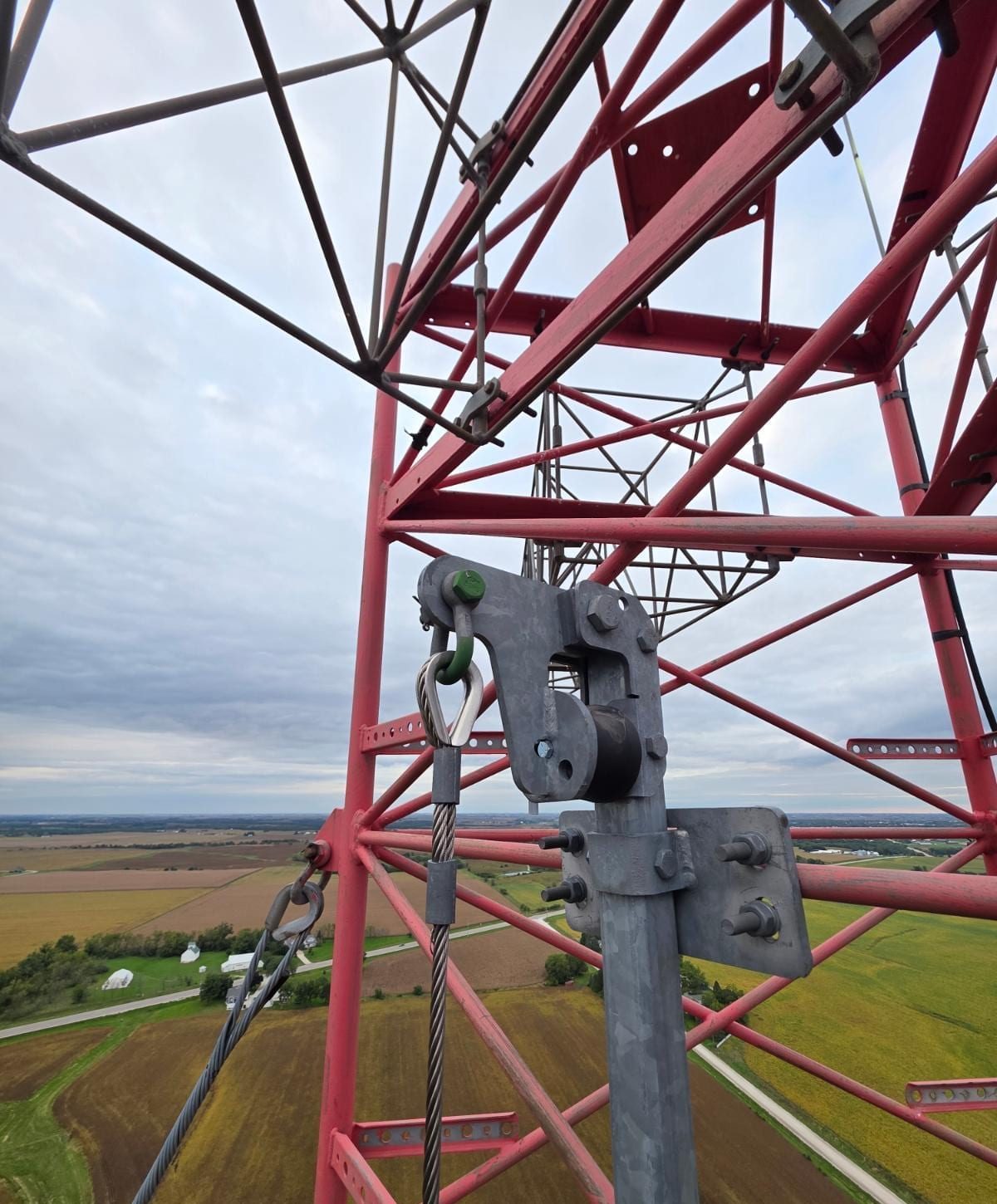 Red lattice tower steel and galvanized cable hardware at the top of the tower, with farmland far below.