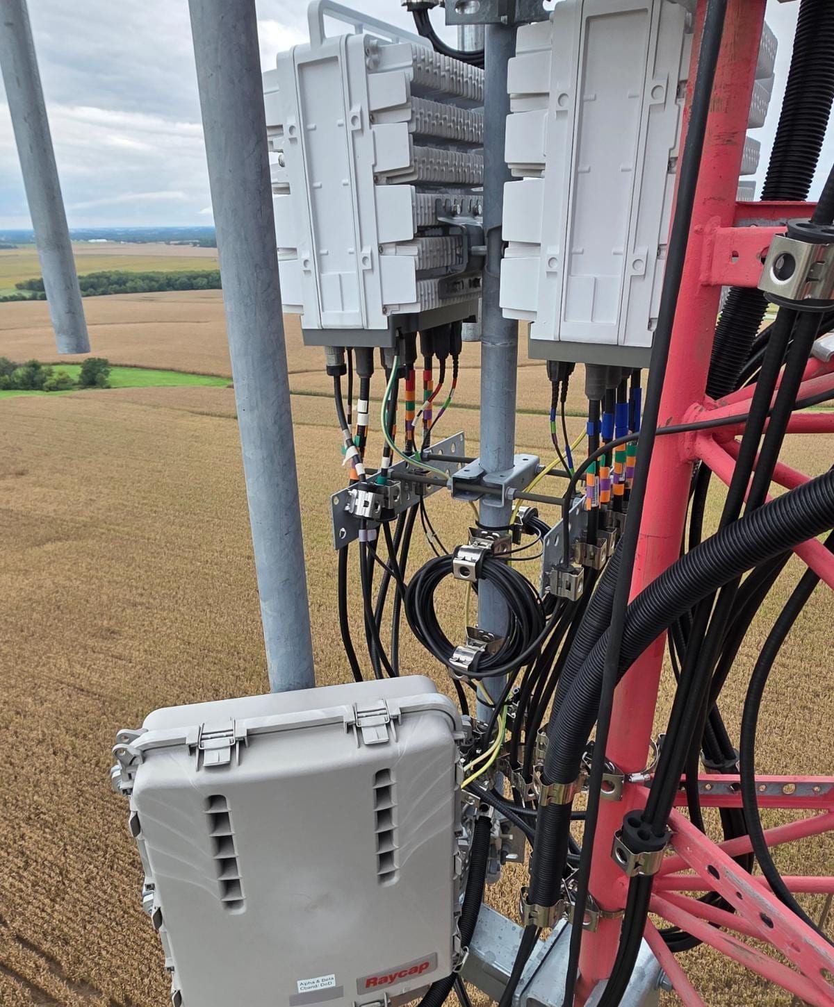 Two radios, a gray junction box and bundled cabling on a tower mount, with fields below.